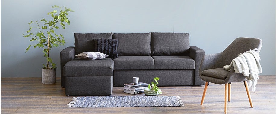 A living room featuring the DELLA sectional sofa bed with storage from JYSK Canada as the centerpiece, providing comfortable seating and storage. The room is decorated with a plant, an accent chair, and a rug, creating a cozy and inviting atmosphere.
