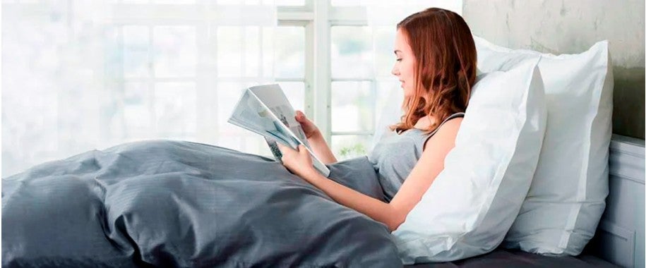 A woman comfortably in bed, propped up by two pillows, engrossed in reading a magazine. 